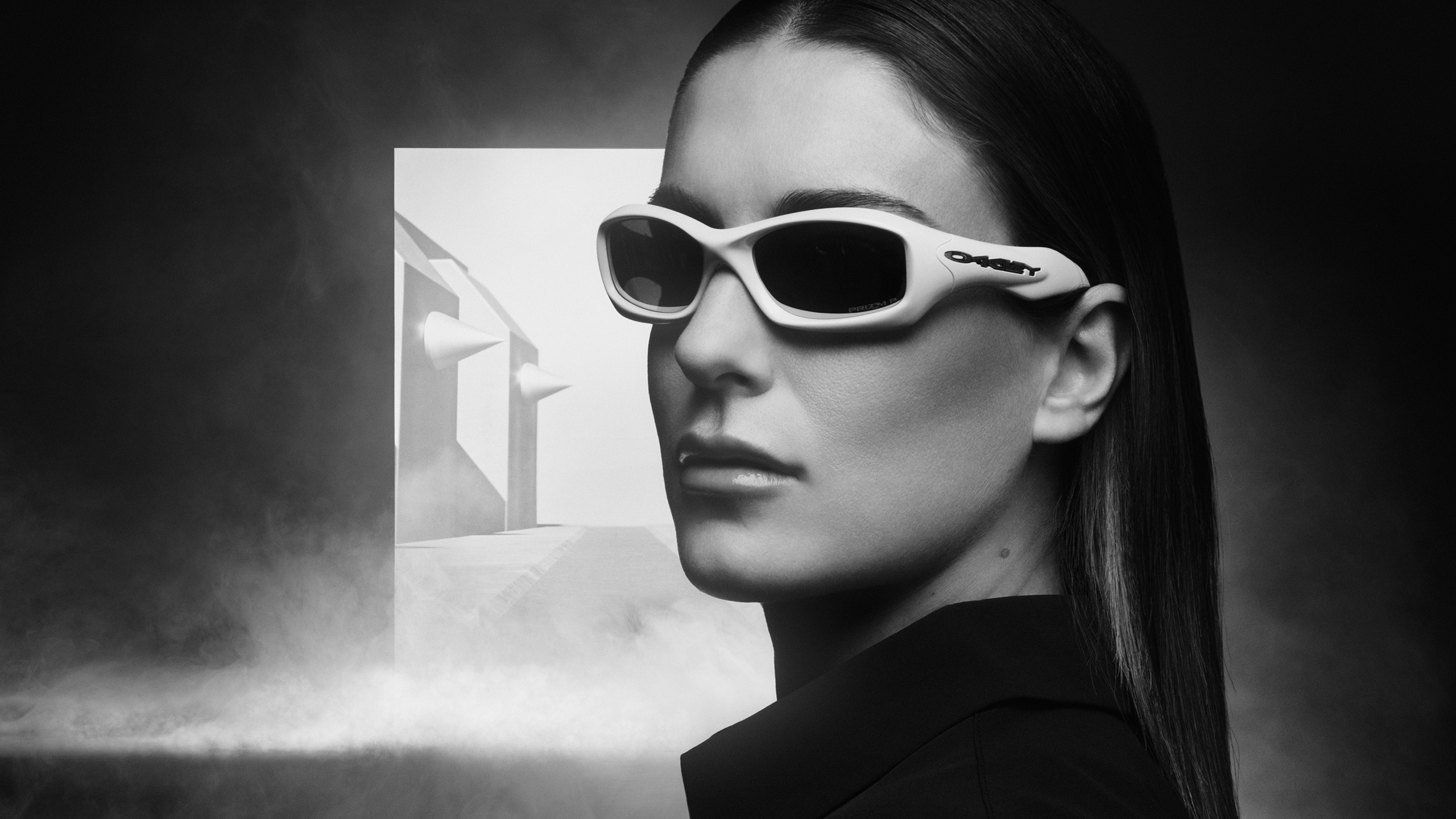 A cinematic, black-and-white portrait of athlete Alexia Putellas wearing light-colored Oakley FatCat sunglasses against a misty, architectural background.
