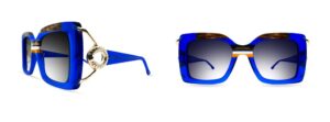 Ultra Limited Torcello sunglasses in electric blue transparent acetate with tortoiseshell and amber colour insertions, gold metal temple featuring a circular jewellery-inspired embellishment with crystal detailing, and gradient blue-grey lenses. Shown in side and front-facing views on white background.