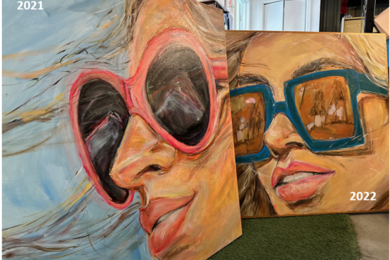 Two large-format oil paintings by artist AnnLo created in collaboration with Transitions®. The 2021 canvas (left) features a close-up portrait of a woman with windswept blonde hair wearing heart-shaped pink sunglasses with dark lenses. The 2022 canvas (right) shows a similar close-up portrait of a woman wearing bold teal square-framed glasses with warm amber-toned reflective lenses, set against a golden ochre background.