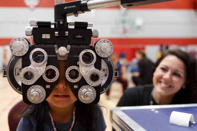 A young student peers through a large, professional phoropter during an eye exam at a school vision clinic. In the background, a LensCrafters volunteer smiles warmly at the child, highlighting the personalized care provided during the event.