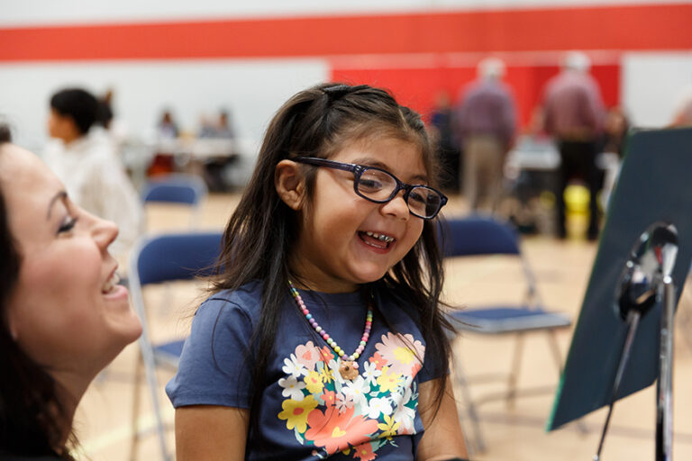Une petite fille portant un t-shirt à fleurs et un collier de perles colorées rit aux éclats en essayant ses nouvelles lunettes de vue à monture noire. Une bénévole est assise à ses côtés, partageant ce moment de joie dans le gymnase transformé en clinique.