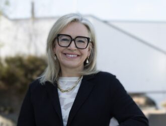 Bronwyn Brophy O'Connor, nouvelle PDG de Carl Zeiss Meditec AG, souriante en extérieur, portant un blazer noir, un haut blanc à motifs, un collier chaîne doré et des lunettes à monture noire, sur fond architectural flou.