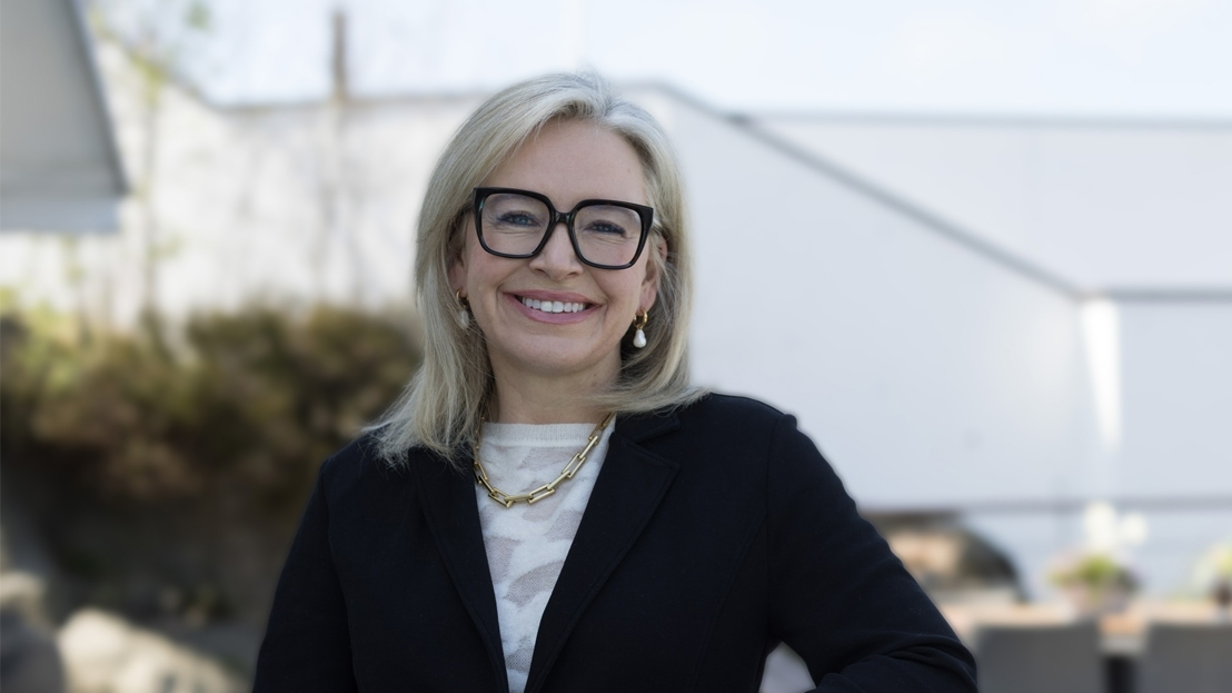 Bronwyn Brophy O'Connor, newly appointed CEO of Carl Zeiss Meditec AG, smiling outdoors in a black blazer, white patterned top, gold chain necklace, and black-rimmed glasses, with a blurred architectural background.
