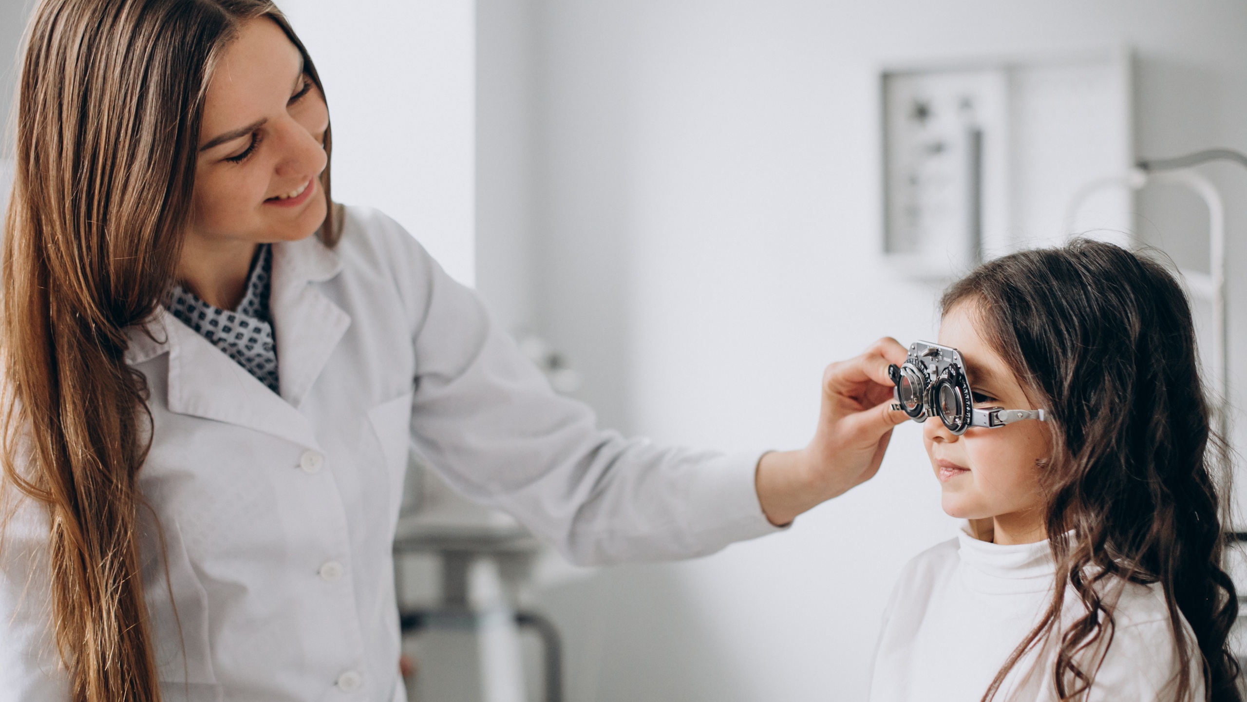 Une opticienne/optométriste en blouse blanche ajustant une monture d'essai (lunettes de test optique) sur une jeune fille lors d'un examen de la vue dans un cadre clinique.