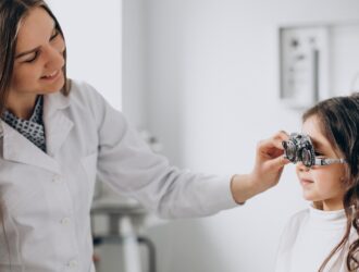 An optometrist in a white lab coat fitting a young girl with a trial frame (optical testing glasses) during an eye examination in a clinical setting.