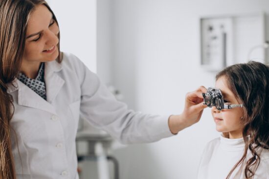 An optometrist in a white lab coat fitting a young girl with a trial frame (optical testing glasses) during an eye examination in a clinical setting.