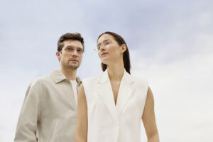 Un homme et une femme portant les lunettes Silhouette The Refined, photographiés en contre-plongée sur un ciel nuageux et doux. Tous deux sont vêtus de tons neutres — l'homme en beige, la femme en blanc — traduisant une esthétique épurée et minimaliste.