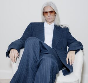 Sarah Harris seated in a cream boucle armchair, wearing oversized gold-framed aviator sunglasses with caramel-tinted lenses, an oversized navy pinstripe suit and white shirt. Her long silver hair falls loosely over her shoulders.