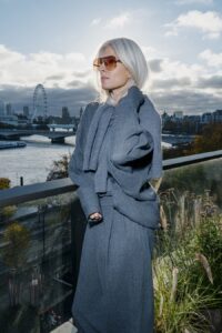 Sarah Harris debout sur une terrasse avec vue sur la Tamise et la Grande Roue de Londres, vêtue d'un manteau en laine gris oversize et d'un tricot assorti, avec des lunettes de soleil aviateur à monture dorée et verres teintés rose. Ses cheveux argentés sont relevés tandis qu'elle regarde au loin.