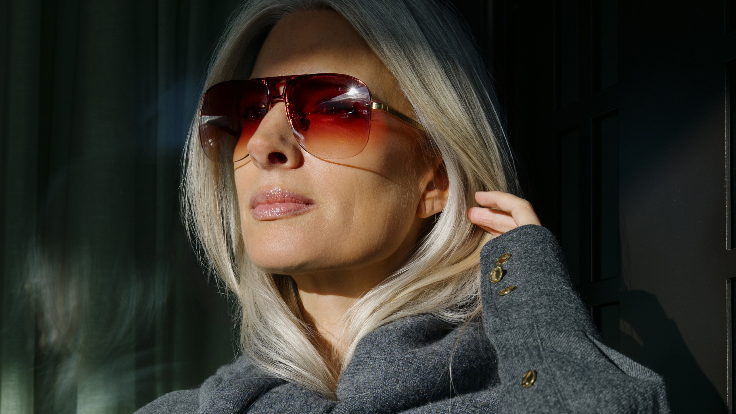 Portrait en gros plan de Sarah Harris portant des lunettes de soleil aviateur sans monture audacieuses avec des verres dégradés rouge rosé intense et une fine monture en titane doré. Elle porte un manteau gris, ses cheveux argentés captant la lumière sur un fond sombre.