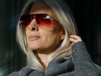 A close-up portrait of Sarah Harris wearing bold rimless aviator sunglasses with deep rose-red gradient lenses and fine gold titanium hardware. She is dressed in a grey coat, her silver hair catching the light against a dark background.