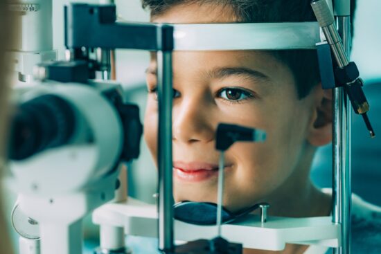 A close-up shot of a young boy receiving an eye exam. His chin and forehead are resting against the supports of a white slit lamp biomicroscope. He is smiling faintly and looking forward as an unseen optometrist, partially visible in the blurry left foreground, conducts the examination.