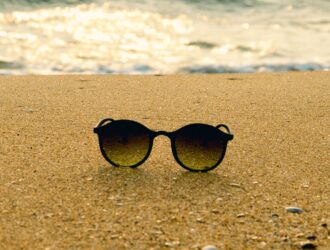 Lunettes de soleil rondes noires sur le sable d'une plage avec l'océan flou en arrière-plan au coucher du soleil.