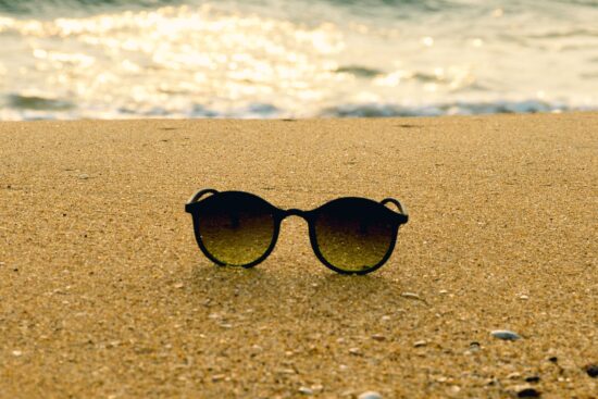 Black round sunglasses on a sandy beach with a blurry ocean background at sunset.
