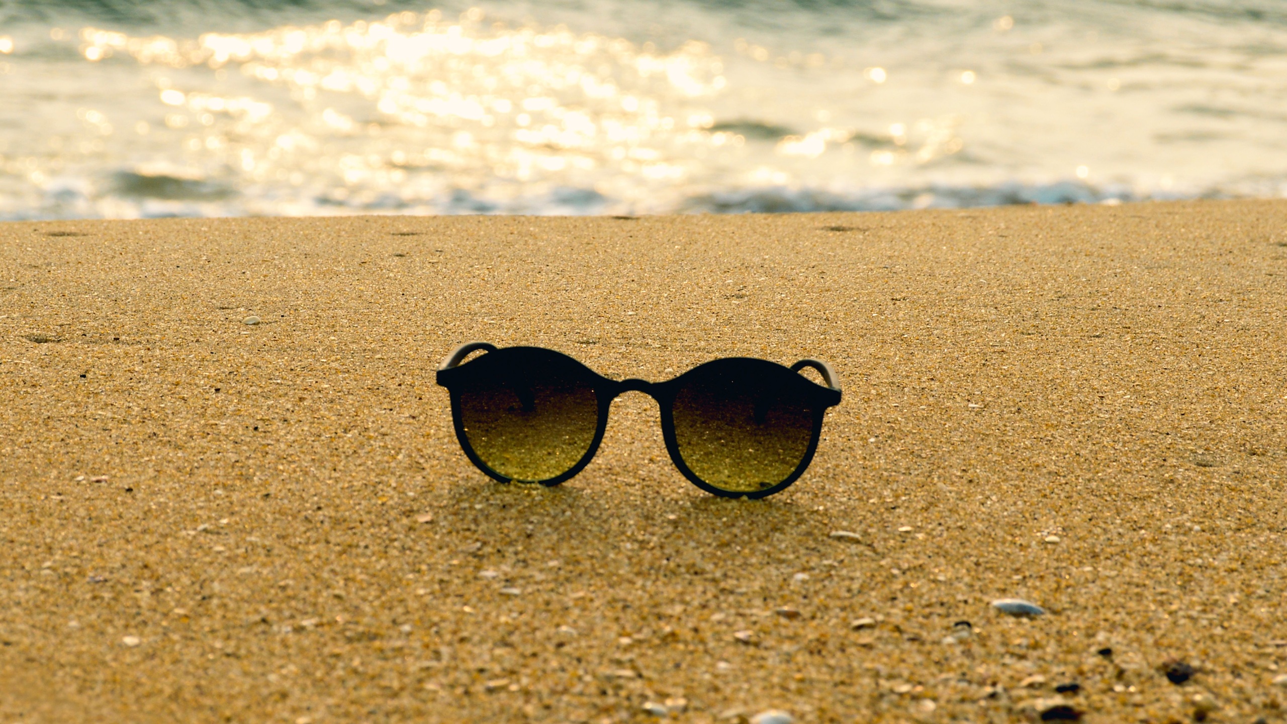 Black round sunglasses on a sandy beach with a blurry ocean background at sunset.