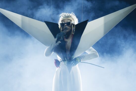 Musical artist Doja Cat performing live on stage during her Tour Ma Vie World Tour, photographed by Greg Noire. Surrounded by dramatic blue stage smoke and bright backlighting, she sings into a microphone while wearing a striking, avant-garde white and black outfit with massive, angular wing-like shoulder structures. She is wearing the custom white and black "Modular Identity" Ray-Ban Meta smart glasses on stage while holding a single red rose.