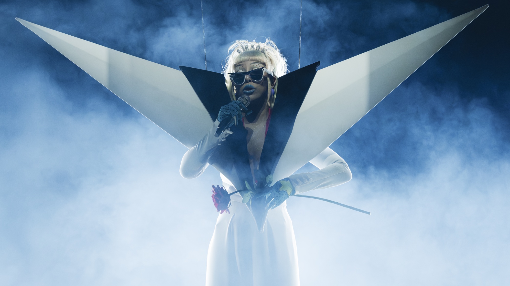 Musical artist Doja Cat performing live on stage during her Tour Ma Vie World Tour, photographed by Greg Noire. Surrounded by dramatic blue stage smoke and bright backlighting, she sings into a microphone while wearing a striking, avant-garde white and black outfit with massive, angular wing-like shoulder structures. She is wearing the custom white and black "Modular Identity" Ray-Ban Meta smart glasses on stage while holding a single red rose.
