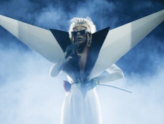 L'artiste Doja Cat en pleine performance sur scène lors de sa tournée mondiale « Tour Ma Vie », photographiée par Greg Noire. Entourée d'une fumée de scène bleue et d'un contre-jour intense, elle chante dans un micro, vêtue d'une tenue blanche et noire avant-gardiste dotée de grandes structures géométriques angulaires aux épaules. Elle porte sur scène les lunettes connectées Ray-Ban Meta personnalisées « Modular Identity » tout en tenant une rose rouge à la main.