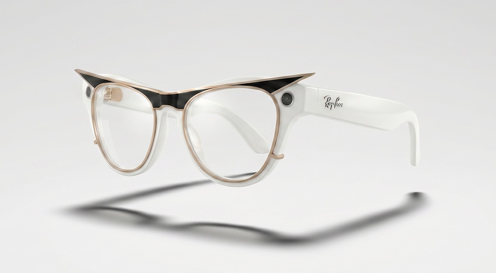 A three-quarter studio product shot of the custom Ray-Ban Meta smart glasses in the "Modular Identity" design, casting a soft shadow on a clean white background. The glasses feature a refined white Skyler frame with clear lenses, enhanced by a black, gold-trimmed sculpted clip-on that dramatically extends the browline into sharp, cat-eye points. The classic Ray-Ban logo is visible in script on the thick white temple arm.