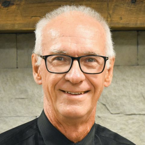 Portrait de face du Dr Roger Jordan souriant, portant des lunettes à monture noire et une chemise noire à col, devant un mur en pierre claire.