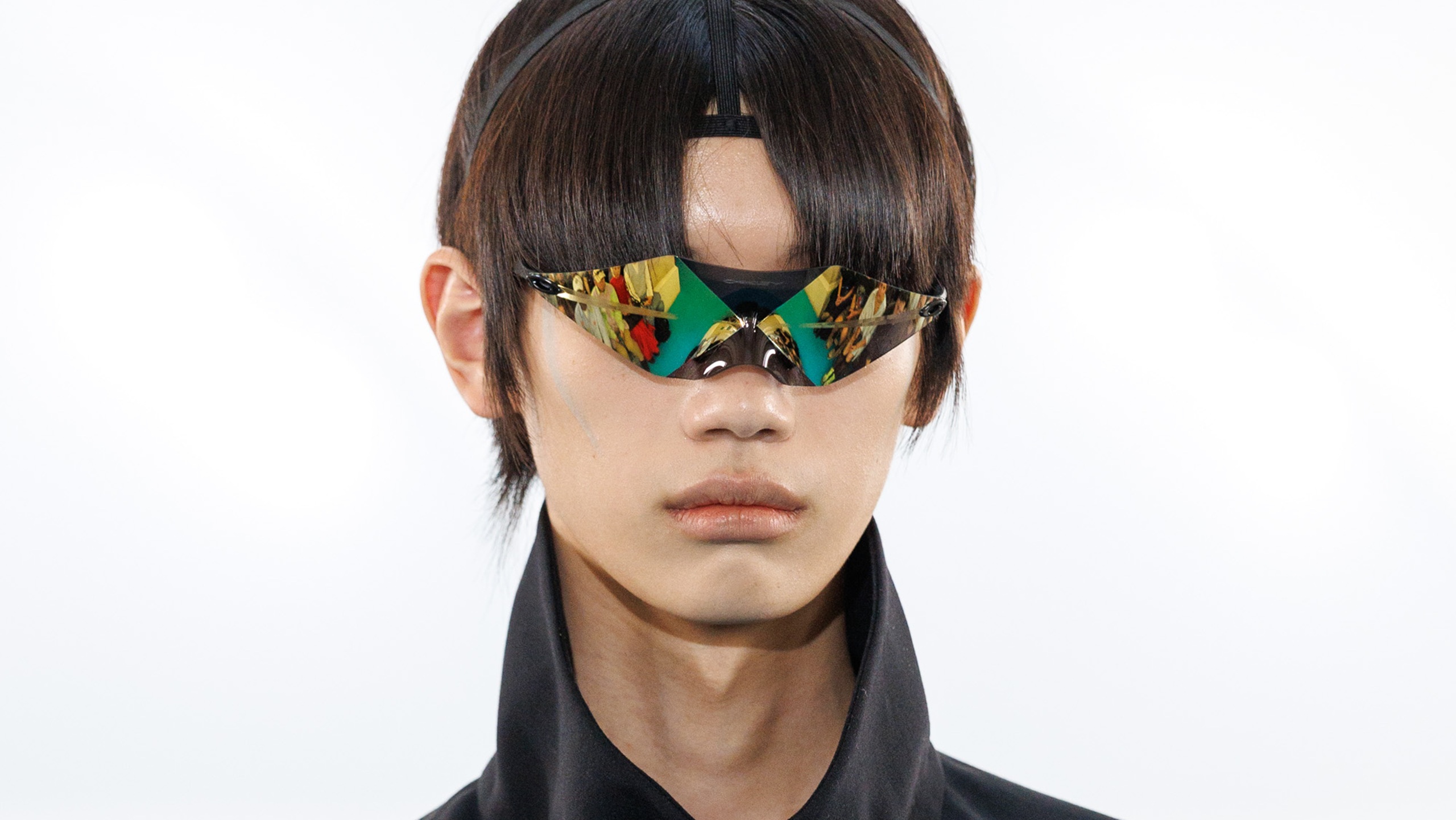 Gros plan sur les lunettes de la collaboration Oakley x Kiko Kostadinov FW26, affichant une silhouette angulaire et des verres réfléchissants aux reflets dorés et verts.