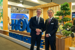 John Goltermann Lassen, CEO of HOYA Vision Care, and Motoki Ozawa, President and CEO of NIDEK, shake hands at the HOYA Vision Care exhibition booth, marking the renewal of their global strategic partnership.