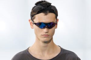 Mannequin du défilé Kiko Kostadinov FW26 portant le prototype de lunettes Oakley au design enveloppant et verres irisés violets et bleus.