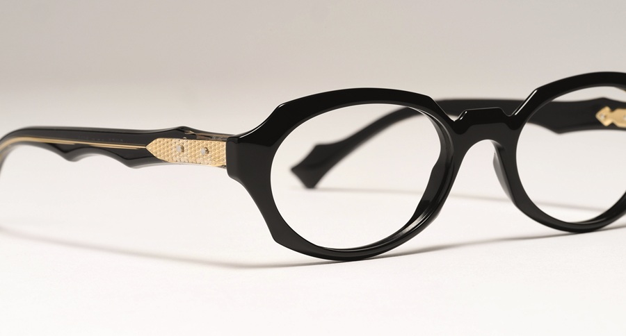 Close-up of black acetate optical glasses featuring a textured gold hinge detail and wave-shaped temples.