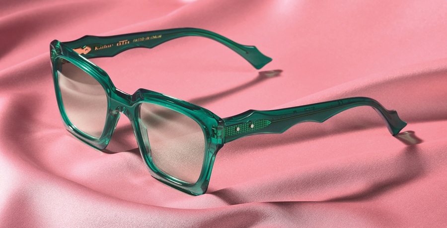Transparent emerald green square sunglasses with wavy temples resting on a draped pink fabric background.