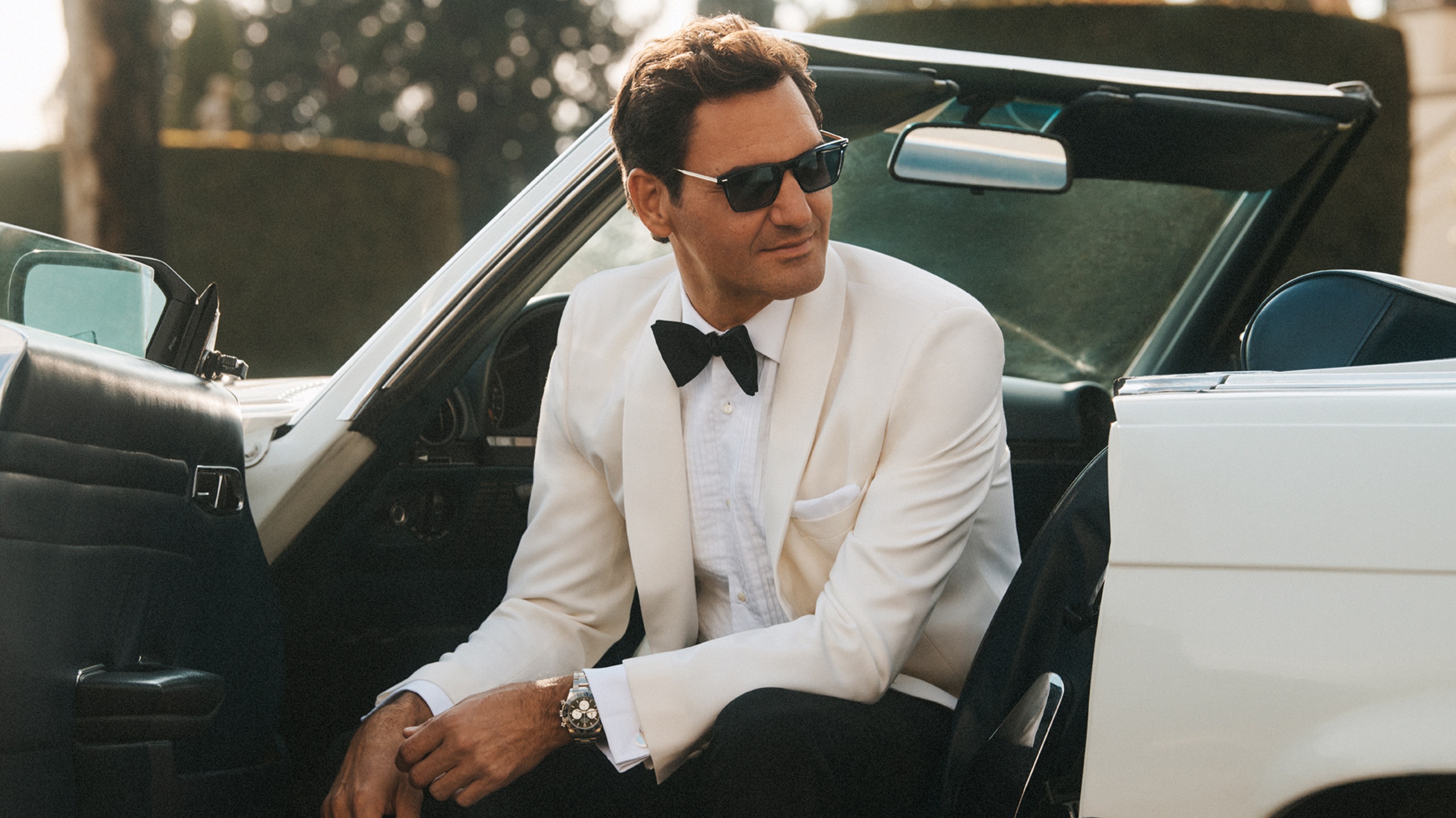 Tennis icon Roger Federer wearing elegant Oliver Peoples sunglasses, dressed in a luxury white tuxedo jacket and black bowtie while sitting in a classic convertible car for the Summer 2026 campaign in Rome.