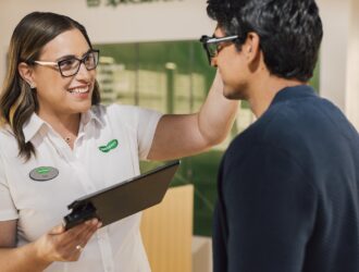 A smiling Specsavers optician adjusts a customer's glasses frames in a Specsavers store, with an eyewear display visible in the background.