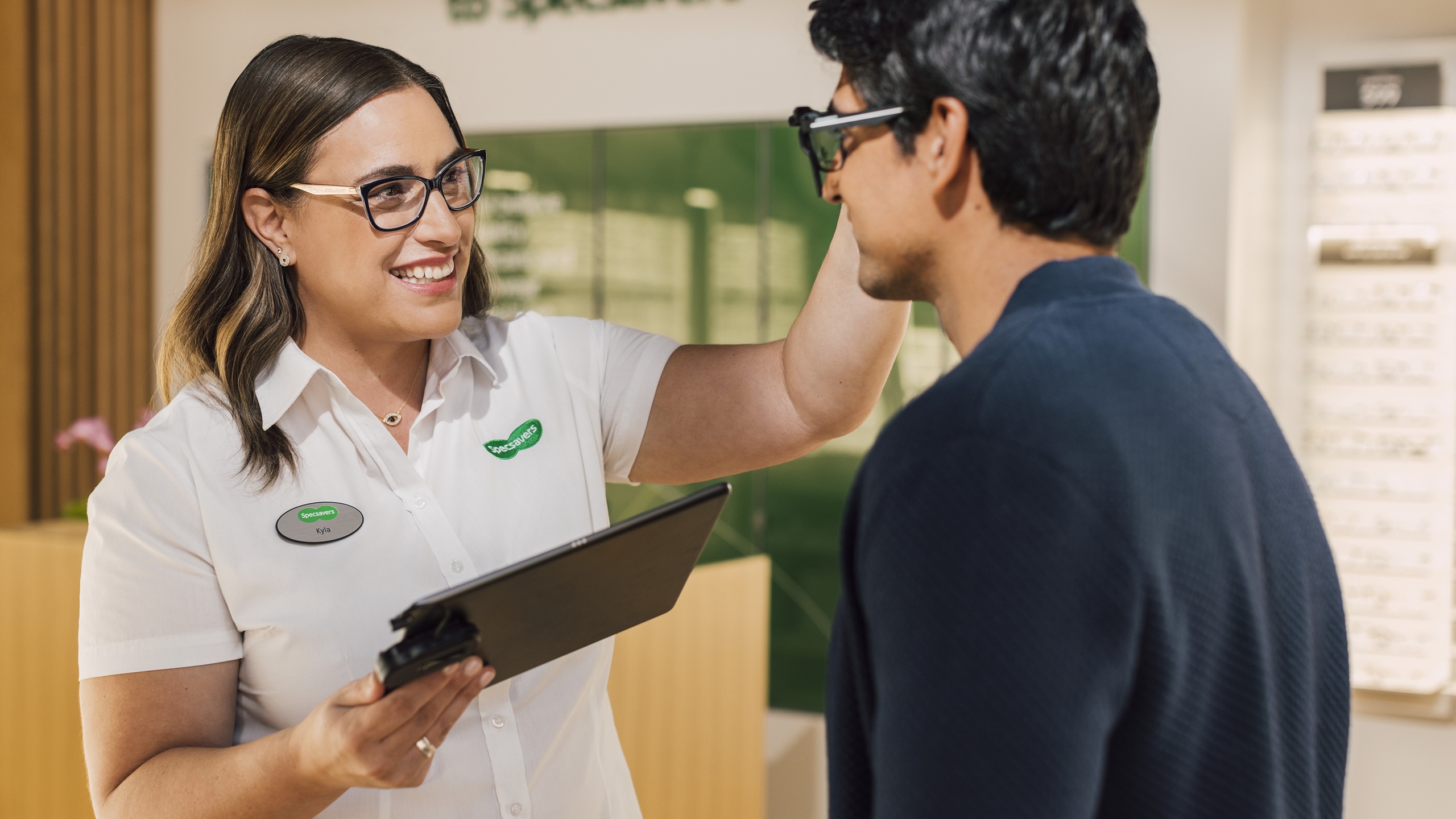 A smiling Specsavers optician adjusts a customer's glasses frames in a Specsavers store, with an eyewear display visible in the background.