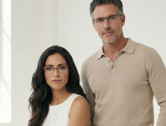 A promotional studio portrait of a woman and a man posing against a bright, minimalist white background next to a window. The woman in the foreground has long dark hair, wearing a white top and purple rectangular titanium eyeglasses. The man standing behind her has short, greying hair and a beard, wearing a beige polo sweater and blue rectangular titanium glasses.