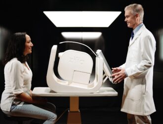 A female patient sitting across a table from a male clinician in a white lab coat who is adjusting the Zilia Ocular, a white, modern retinal imaging device placed on the table between them.