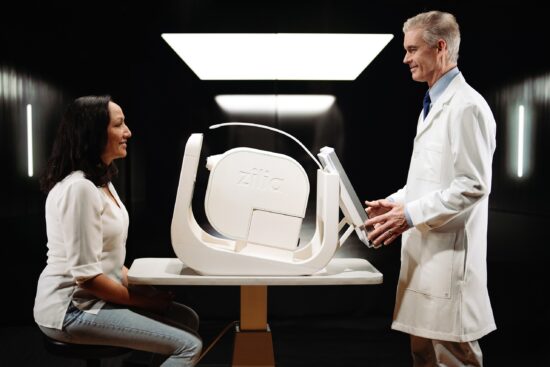 A female patient sitting across a table from a male clinician in a white lab coat who is adjusting the Zilia Ocular, a white, modern retinal imaging device placed on the table between them.