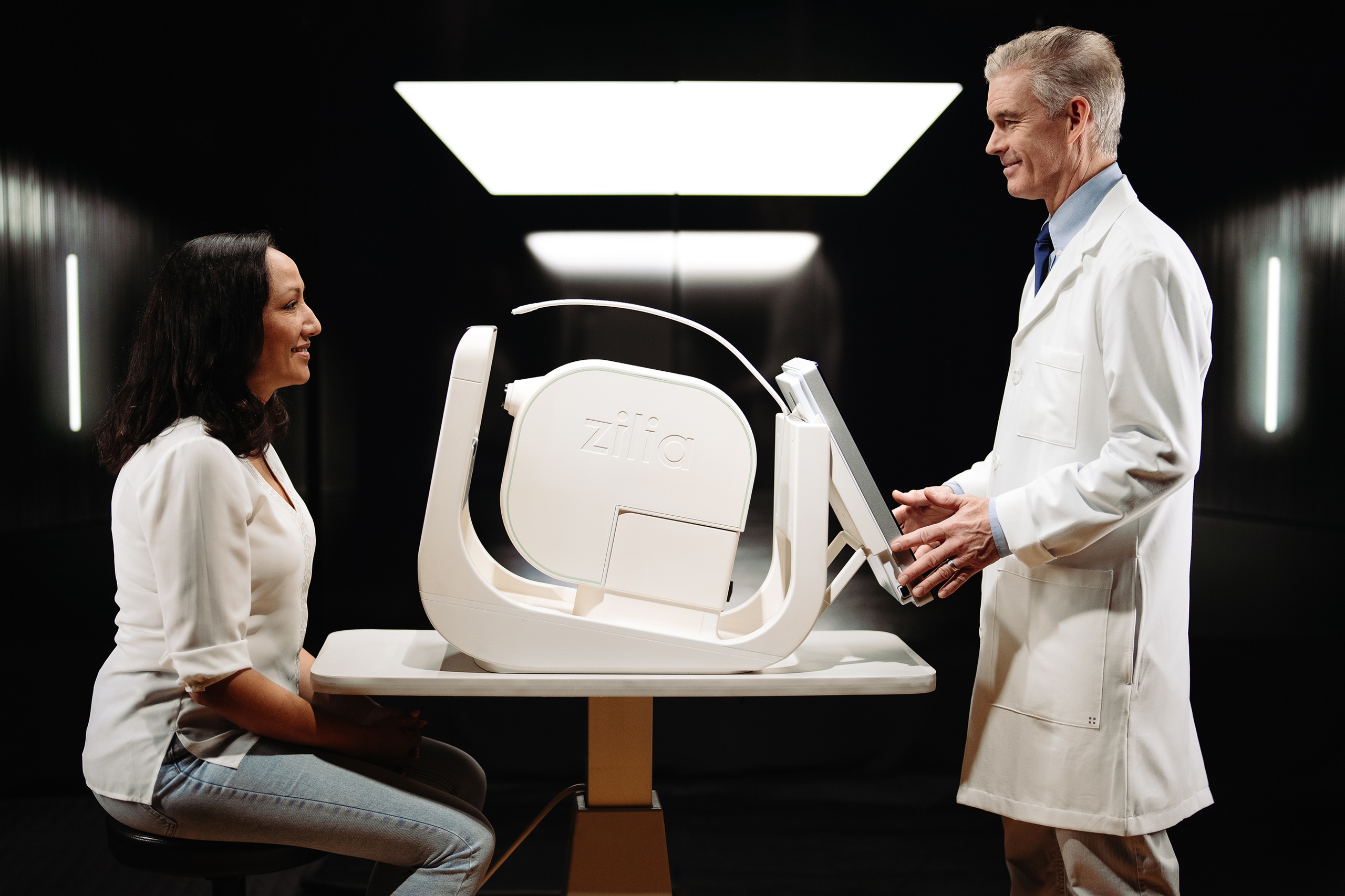 A female patient sitting across a table from a male clinician in a white lab coat who is adjusting the Zilia Ocular, a white, modern retinal imaging device placed on the table between them.