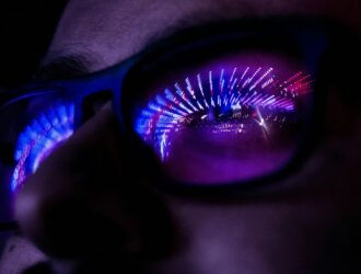 Gros plan d’une personne portant des lunettes à monture foncée dans une ambiance sombre. Des lumières néon violettes, bleues et roses se reflètent dans les verres, formant un motif abstrait en forme de tunnel et éclairant une partie du visage.