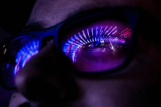 Close-up of a person wearing dark-framed glasses in low light. Bright purple, blue, and pink neon lights are reflected in the lenses, creating an abstract tunnel-like pattern and illuminating part of the face.