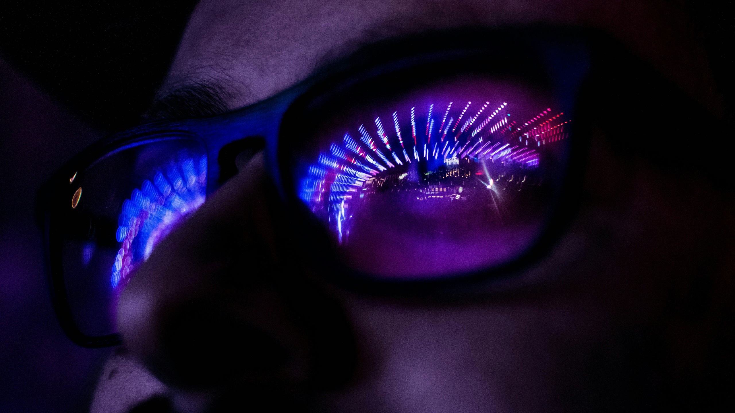 Close-up of a person wearing dark-framed glasses in low light. Bright purple, blue, and pink neon lights are reflected in the lenses, creating an abstract tunnel-like pattern and illuminating part of the face.