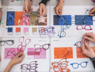 Top-down view of hands arranging colorful titanium eyewear frames over design mood boards and Pantone color swatches on a table.