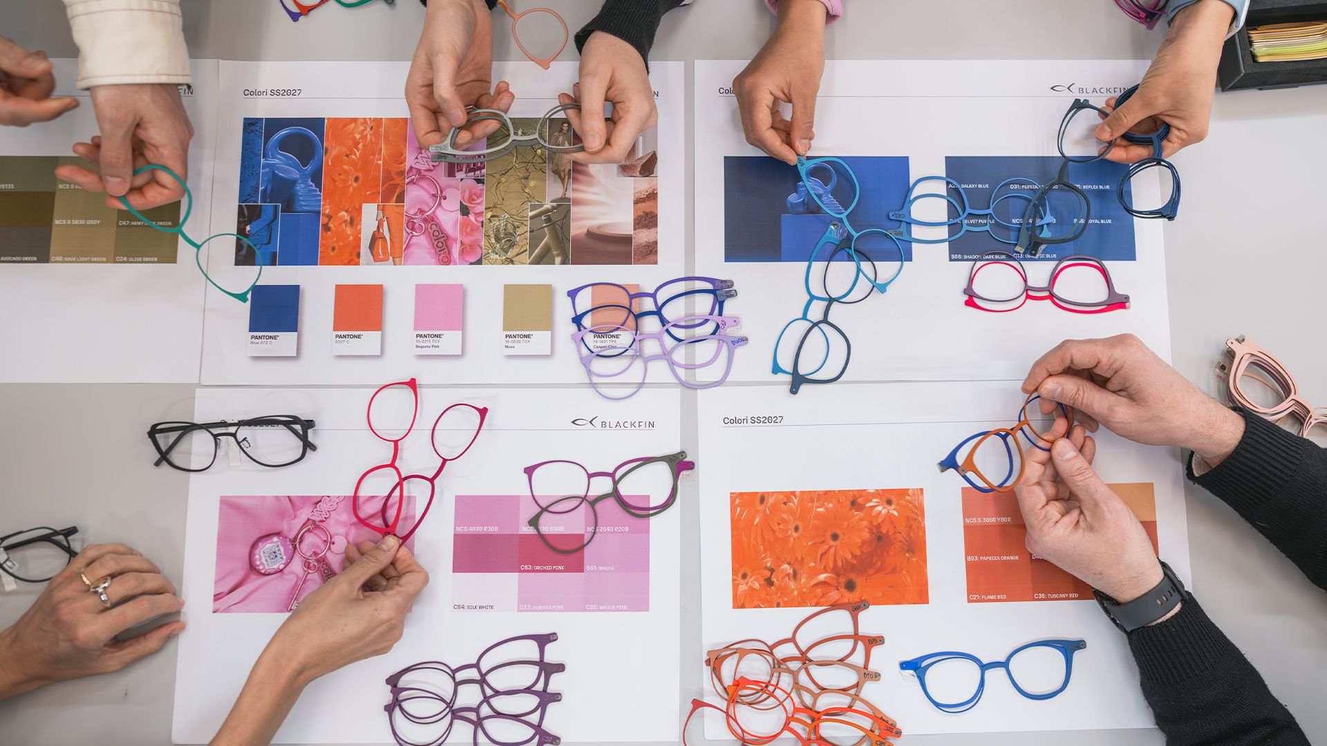Top-down view of hands arranging colorful titanium eyewear frames over design mood boards and Pantone color swatches on a table.