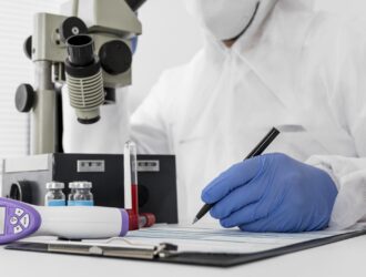 Gros plan d'un professionnel de la santé ou chercheur en combinaison de protection blanche, masque et gants en nitrile bleus, écrivant sur un presse-papiers. En arrière-plan, un grand microscope de laboratoire côtoie de petits flacons médicaux et un tube à essai. Un thermomètre infrarouge numérique est posé sur le bureau au premier plan.