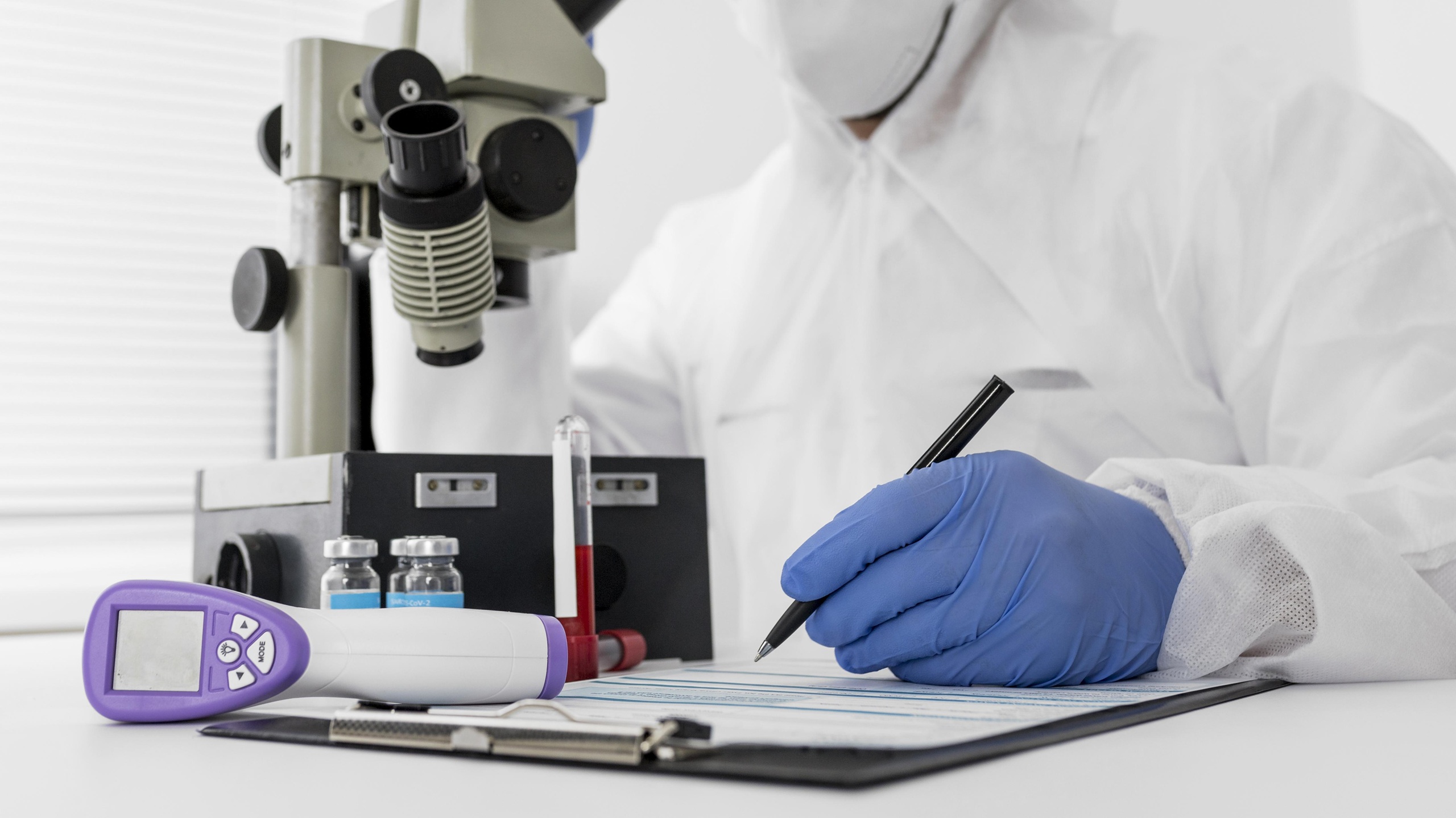 Gros plan d'un professionnel de la santé ou chercheur en combinaison de protection blanche, masque et gants en nitrile bleus, écrivant sur un presse-papiers. En arrière-plan, un grand microscope de laboratoire côtoie de petits flacons médicaux et un tube à essai. Un thermomètre infrarouge numérique est posé sur le bureau au premier plan.