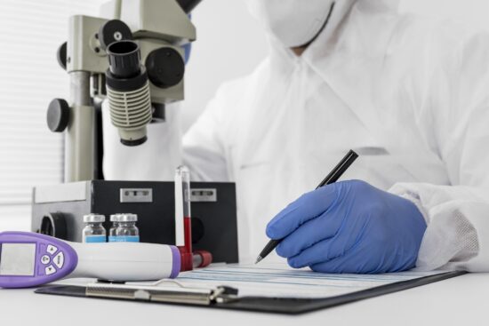 Close-up of a healthcare professional or researcher in a white hazmat suit, face mask, and blue nitrile gloves writing on a clipboard. In the background, a large laboratory microscope sits next to small medical vials and a test tube. A digital infrared thermometer rests on the desk in the foreground.