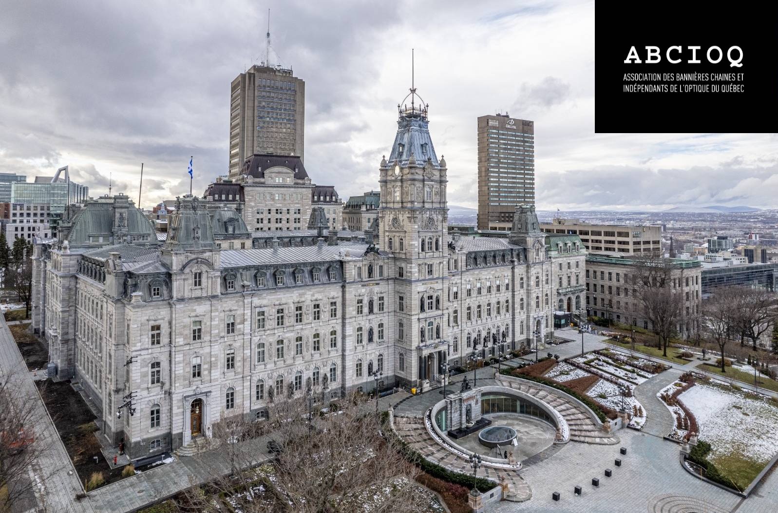 Vue aérienne de l'Hôtel du Parlement de Québec, un imposant édifice de style Second Empire en pierre grise, entouré de ses jardins en hiver avec des traces de neige. Des gratte-ciels modernes se dressent en arrière-plan sous un ciel nuageux. Le logo de l'ABCIOQ apparaît dans un encadré noir en haut à droite de l'image.