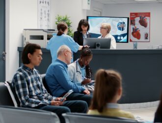 A busy medical clinic waiting room and reception area. In the foreground, patients sit in a row of black chairs, including a young man in a plaid shirt and an older man in a blue shirt talking to a female doctor with a stethoscope. Behind them, a curved dark grey reception desk features a female receptionist, a doctor in a white coat, and a nurse in blue scrubs looking at a computer screen. The background includes medical posters, a large monitor, and a water dispenser