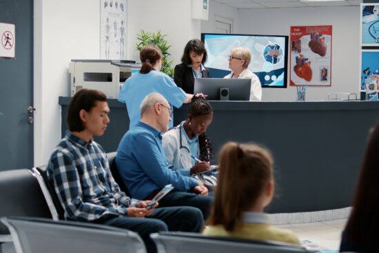 A busy medical clinic waiting room and reception area. In the foreground, patients sit in a row of black chairs, including a young man in a plaid shirt and an older man in a blue shirt talking to a female doctor with a stethoscope. Behind them, a curved dark grey reception desk features a female receptionist, a doctor in a white coat, and a nurse in blue scrubs looking at a computer screen. The background includes medical posters, a large monitor, and a water dispenser