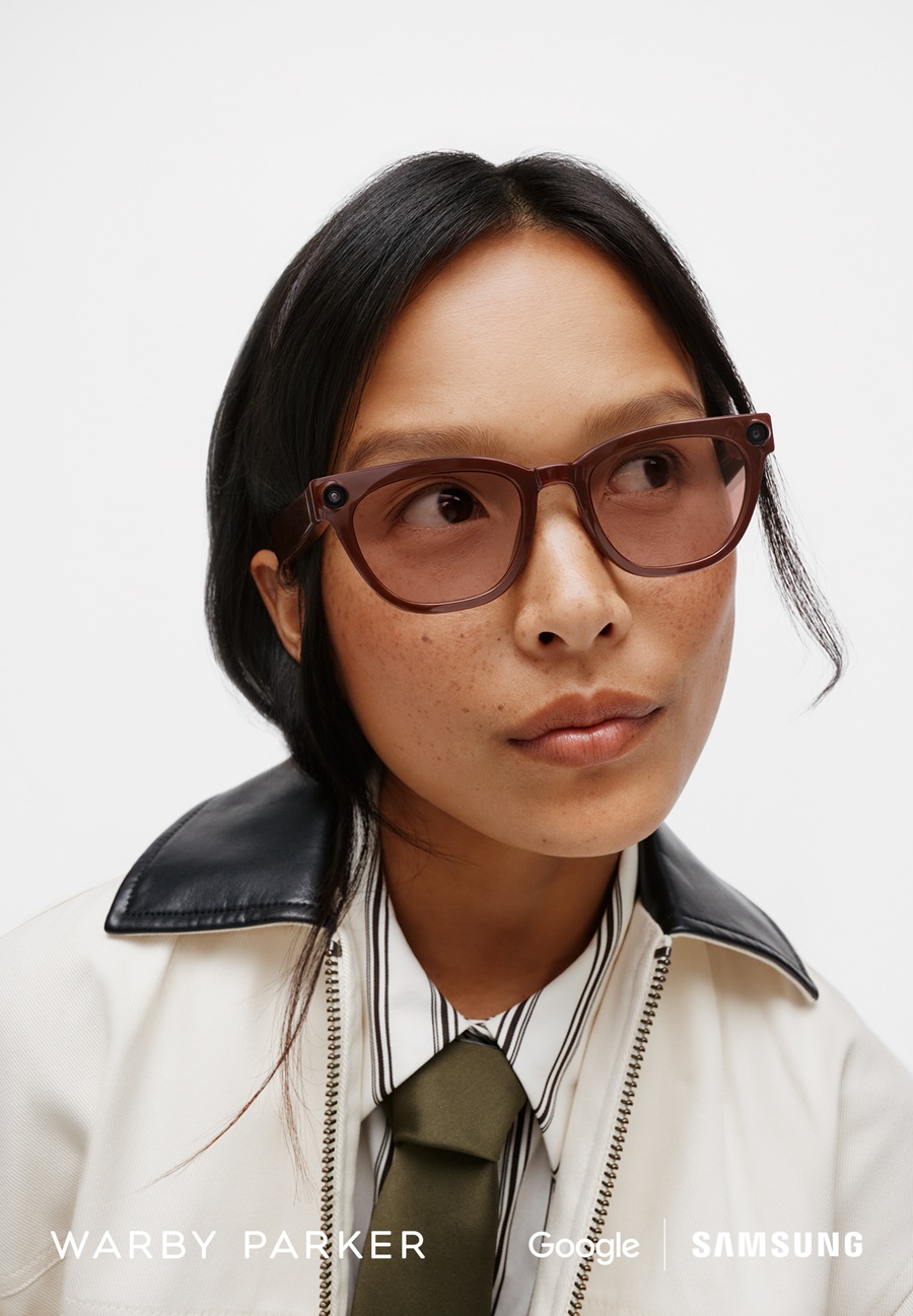 Close-up lifestyle portrait of a female model wearing the amber Samsung Intelligent Eyewear (Warby Parker design) with brown-tinted lenses. She is wearing a cream jacket with a black collar. Logos for Warby Parker, Google, and Samsung are visible.