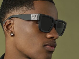 Close-up profile of a man wearing black square-frame True Religion sunglasses featuring the brand's horseshoe logo on the temple.