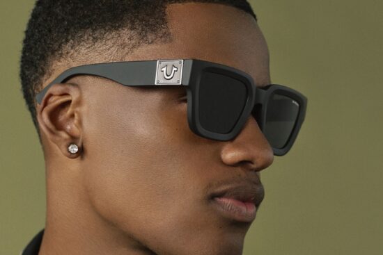 Close-up profile of a man wearing black square-frame True Religion sunglasses featuring the brand's horseshoe logo on the temple.